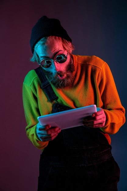 Foto grátis usando o tablet, parece chocado. retrato do homem caucasiano em fundo gradiente de estúdio em luz de néon. lindo modelo masculino com estilo hippie. conceito de emoções humanas, expressão facial, vendas, anúncio.