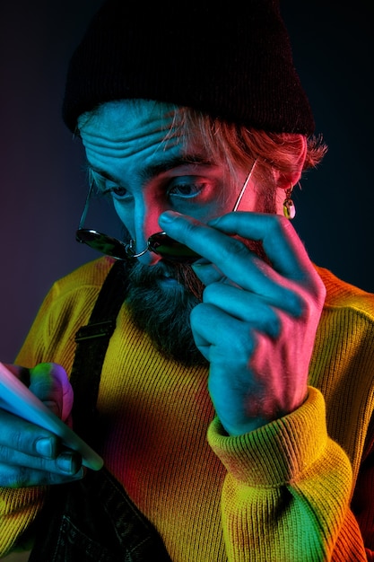 Usando o telefone em óculos de sol. Retrato do homem caucasiano em fundo gradiente de estúdio em luz de néon. Lindo modelo masculino com estilo hippie. Conceito de emoções humanas, expressão facial, vendas, anúncio.