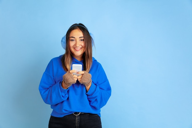 Foto grátis usando o telefone, feliz. retrato de uma mulher caucasiana sobre fundo azul do estúdio. linda modelo feminino com roupas quentes. conceito de emoções humanas, expressão facial, vendas, anúncio. clima de inverno, feriados.