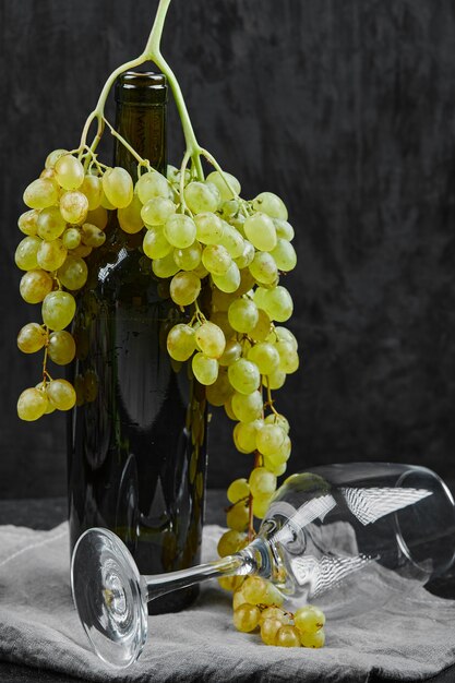 Uvas brancas em torno de uma garrafa de vinho e um copo vazio em fundo escuro. Foto de alta qualidade