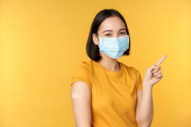 Vacinação do conceito de covid e saúde Imagem de menina coreana sorridente em curativo de máscara facial médica no ombro, apontando o dedo para o fundo amarelo de anúncio de banner