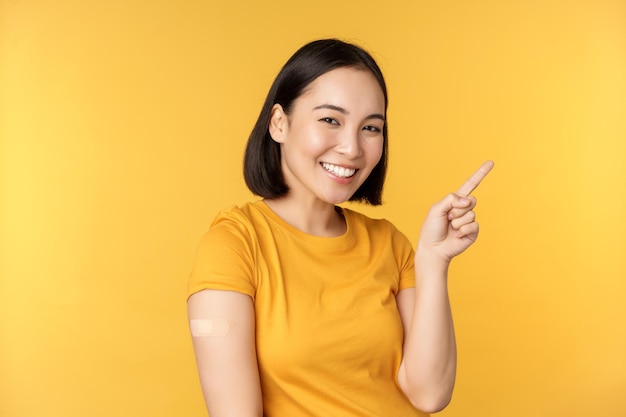 Vacinação e conceito de pandemia covid19 Mulher coreana sorridente com band-aid no ombro após vacina contra coronavírus disparada apontando para banner com campanha de vacinação