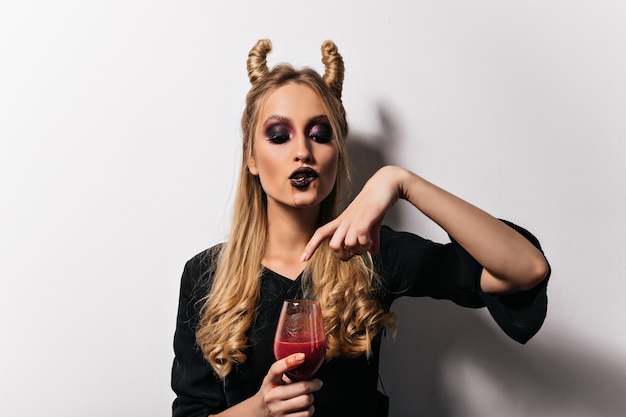 Vampira bebendo sangue de um copo de vinho. Bela bruxa loira desfrutando de poição no halloween.