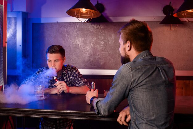 Vaping homem segurando um mod. Uma nuvem de vapor no vapebar. Dois homens descansam em um bar e fumam cigarros eletrônicos.