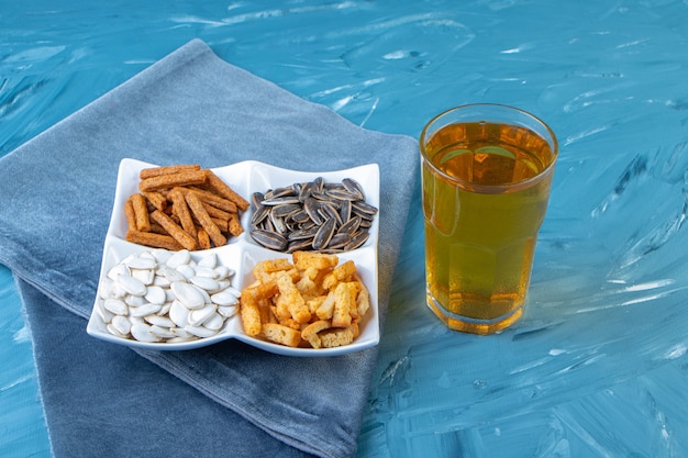 Várias tigelas de lanche ao lado do copo de cerveja em uma toalha, na superfície azul.
