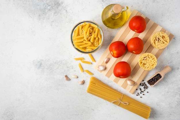 Variedade de massa crua, garrafa de azeite, grãos de pimenta e tomate em fundo branco.