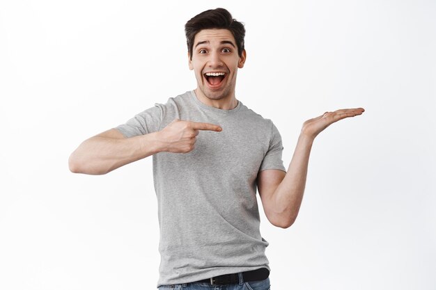 Veja isso. Homem animado e feliz apontando para a mão com seu produto, mostrando o item na palma da mão contra o espaço da cópia, demonstrar logotipo, fundo branco