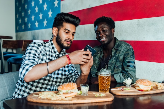 Velhos amigos alegres se divertindo com o smartphone e bebendo chope com hambúrgueres no balcão do bar no pub.