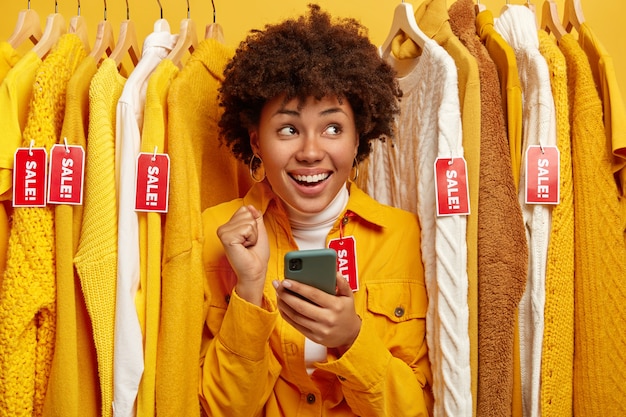 Venda, moda, descontos e conceito de compras online. Mulher de pele escura muito feliz escolhe roupas em loja de roupas, alegra grandes vendas, mantém o celular