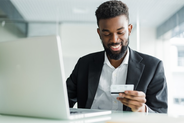 Vendas bancárias na Internet. Empresário africano bem sucedido, sentado em um laptop e segurando o cartão de crédito na mão até o empresário fazer pedidos pela Internet