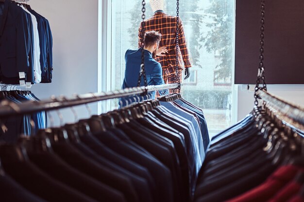 Vendedor barbudo elegante se preocupa com terno em um manequim em uma loja de moda masculina.