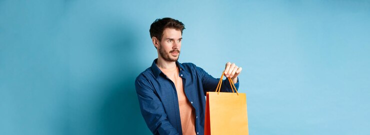 Foto grátis vendedor de compras jovem bonito estendendo a sacola de compras laranja para o cliente olhando de lado stan