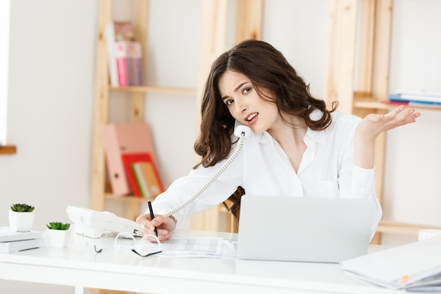 Vendedora séria e bem-vestida falando ao telefone no escritório atrás de sua mesa e copiando o espaço do laptop