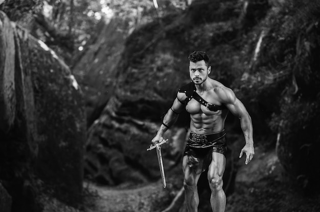 Foto grátis venha até mim. foto monocromática de um jovem gladiador masculino segurando uma espada pronta para lutar perto das rochas