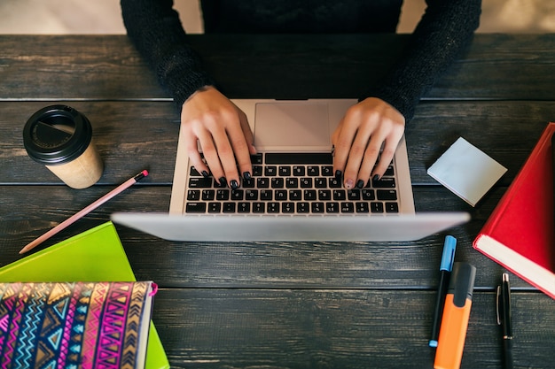 Ver os de cima nas mãos de uma linda mulher sentada à mesa na camisa preta, trabalhando no laptop em co-working office, artigos de papelaria, mãos digitando, bebendo café, freelance