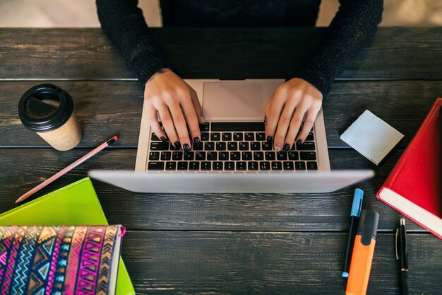 Ver os de cima nas mãos de uma linda mulher sentada à mesa na camisa preta, trabalhando no laptop em co-working office, artigos de papelaria, mãos digitando, bebendo café, freelance