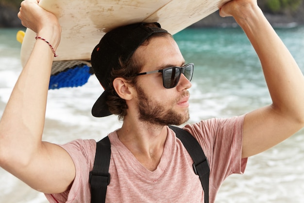 Verão, férias, lazer e conceito de estilo de vida ativo. Na moda jovem turista vestindo snapback e óculos de sol em pé na beira-mar e olhando para a distância