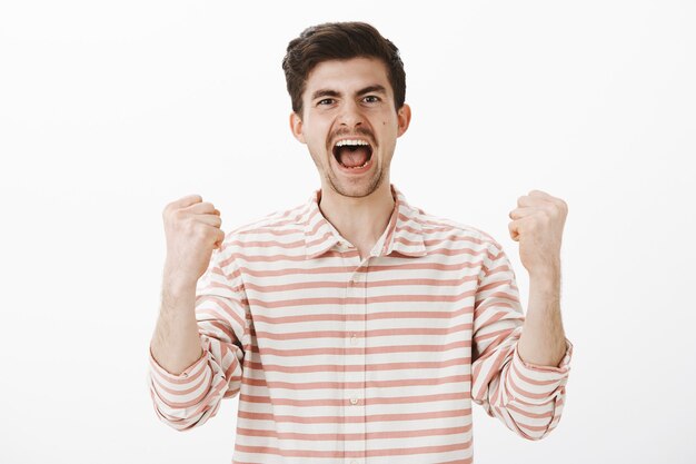 Verdadeiro torcedor alegre por seu time favorito. Foto de um belo colega de trabalho triunfando, gritando de felicidade e vitória, erguendo os punhos cerrados, celebrando a vitória, sentindo-se como um campeão sobre a parede cinza