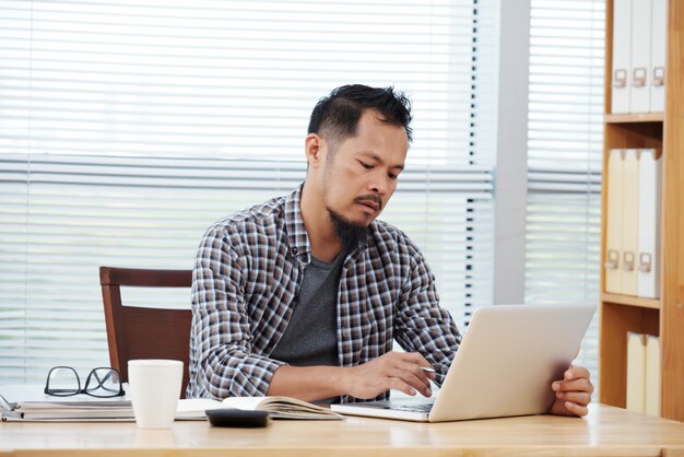 Vestido casualmente homem filipino sentado no escritório e trabalhando no laptop