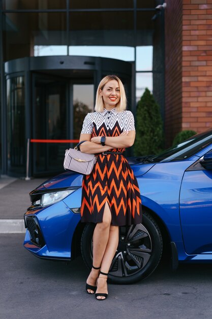 Vestido de mulher jovem e deslumbrante posando na frente de seu carro ao ar livre, motorista de propriedade