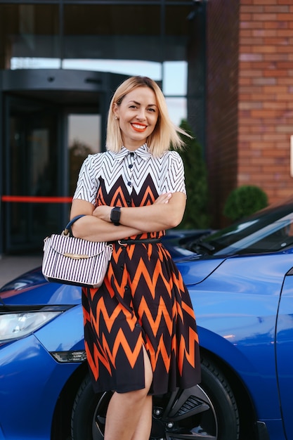 Vestido de mulher jovem e deslumbrante posando na frente de seu carro ao ar livre, motorista de propriedade