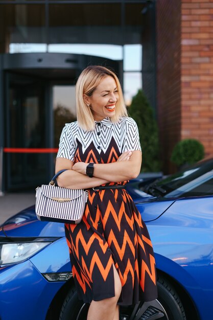 Vestido de mulher jovem e deslumbrante posando na frente de seu carro ao ar livre, motorista de propriedade