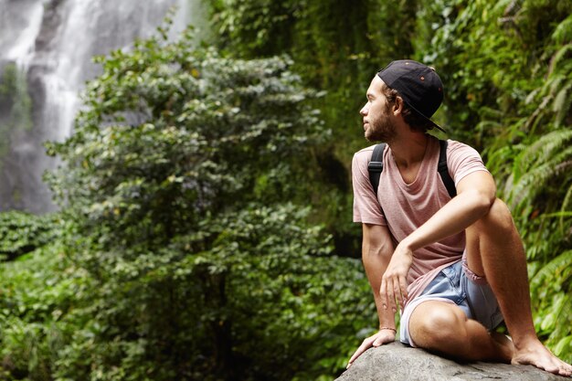 Viagem e aventura. Jovem bonito caminhante descalço com uma mochila relaxando sozinho em uma pedra grande e olhando para trás