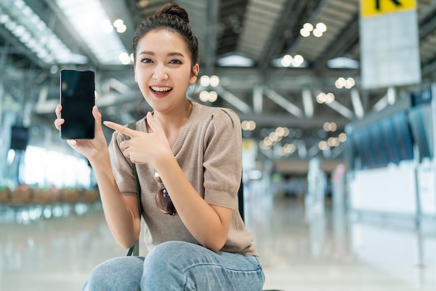 Viagem segurajovem atraente sorridente mulher asiática mão mostrar smartphone de tela em branco com felicidade alegre viagem férias após o bloqueio é superasiática feminina sente-se relaxe no terminal do aeroporto