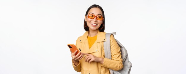 Viajante de turista elegante jovem asiática com mochila e smartphone sorrindo para a câmera posando contra fundo branco
