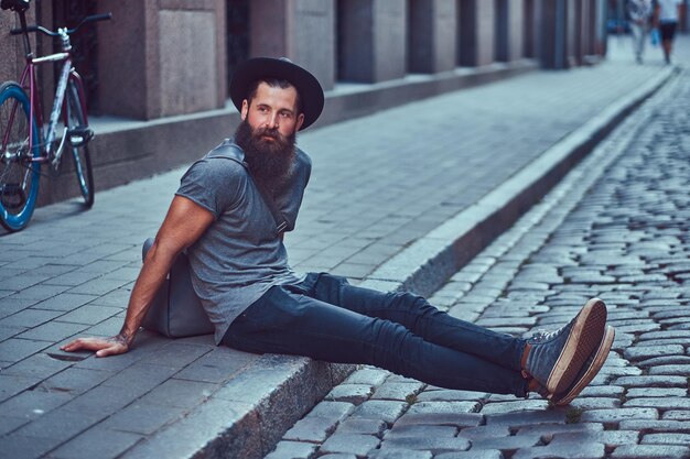 Viajante hipster bonito com uma barba elegante e tatuagem nos braços, vestido com roupas casuais com uma bolsa, senta-se na calçada, descansando depois de um passeio de bicicleta.