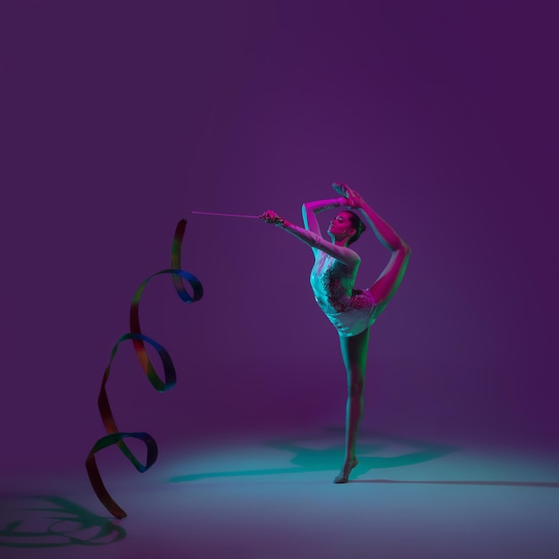 Vibrante. Jovem atleta feminina, artista de ginástica rítmica dançando, treinando com fita isolada no fundo roxo do estúdio na luz de neon. Linda garota praticando com equipamento. Graça no desempenho