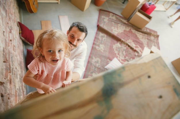 Vida nova. O jovem pai e a filha mudaram-se para uma nova casa ou apartamento. Pareça feliz e confiante. Movimento, relações, conceito de estilo de vida. Brincando juntos, preparando-se para consertar e rindo.