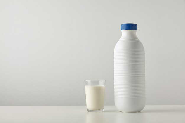 Vidro transparente com leite orgânico fresco perto de uma garrafa de plástico folheada em branco com tampa azul isolada na lateral da mesa branca.