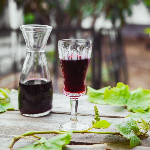 Vinho tinto no jarro e copo com vista lateral de galho de árvore de uva na mesa de madeira e jardim