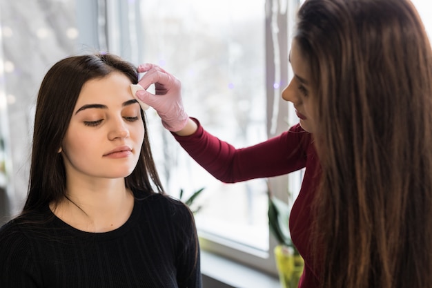 Visagiste hábil, fazendo sobrancelhas, maquiagem para uma jovem mulher com cabelos escuros