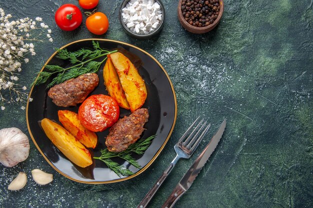 Visão aérea de deliciosas costeletas de carne assadas com batatas e tomates em uma placa preta talheres com flores brancas especiarias alhos no fundo verde preto cores misturadas