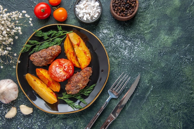 Visão aérea de deliciosas costeletas de carne assadas com batatas e tomates em uma placa preta talheres com flores brancas especiarias alhos no fundo verde preto cores misturadas