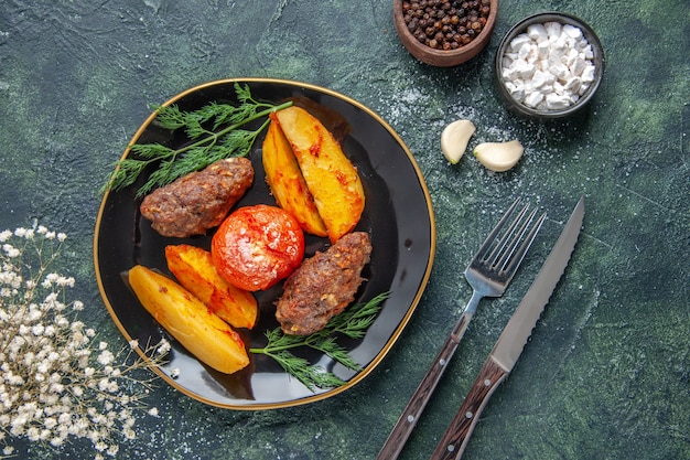 Visão aérea de deliciosas costeletas de carne assadas com batatas e tomates em uma placa preta talheres de especiarias de alhos em um fundo de cores preto e verde