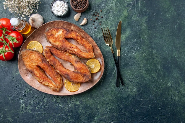 Visão aérea de deliciosos peixes fritos e rodelas de limão em um prato marrom temperos com tomate e garrafa de óleo na mesa de cores com espaço livre