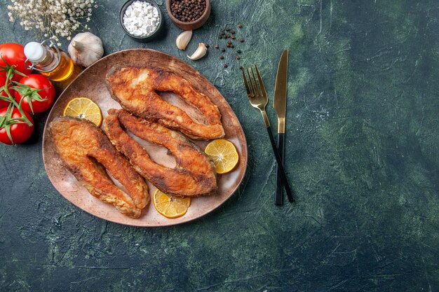 Visão aérea de deliciosos peixes fritos e rodelas de limão em um prato marrom temperos com tomate e garrafa de óleo na mesa de cores com espaço livre