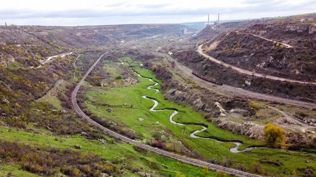 Visão aérea de drones da natureza na Moldávia, fluxo do riacho que flui para a ravina, encostas com vegetação esparsa e rochas, trem em movimento, céu nublado