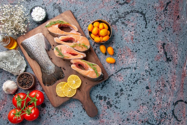 Visão aérea de peixes crus rodelas de limão verdes pimenta no lado direito na tábua de madeira e flores na mesa de cores preto azul