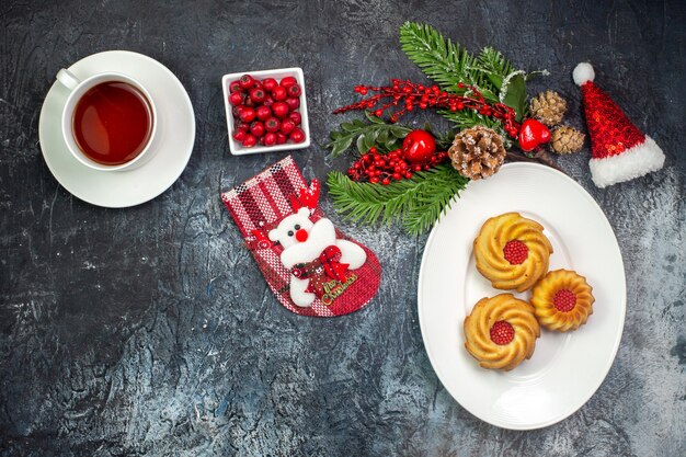 Visão aérea de uma xícara de chá com biscoitos deliciosos em um prato branco, chapéu de Papai Noel e chocolate em uma tigela na superfície escura