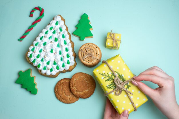 Visão aérea de vários biscoitos de açúcar da árvore de Natal e a mão segurando uma caixa de presente amarela sobre fundo azul pastel