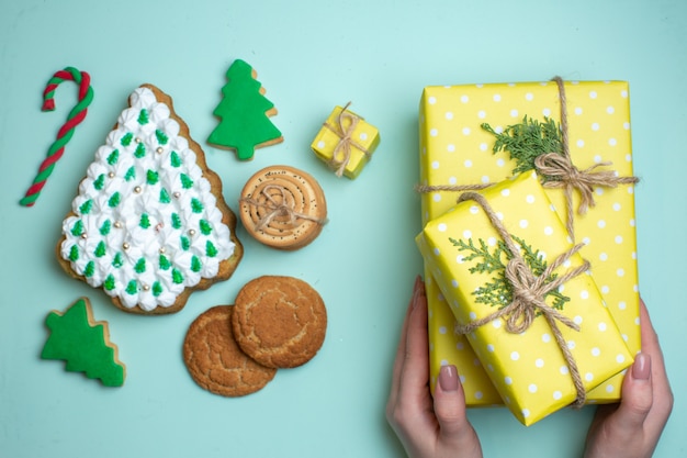 Visão aérea de vários biscoitos de açúcar da árvore de natal e a mão segurando uma das caixas de presente amarela sobre fundo azul pastel