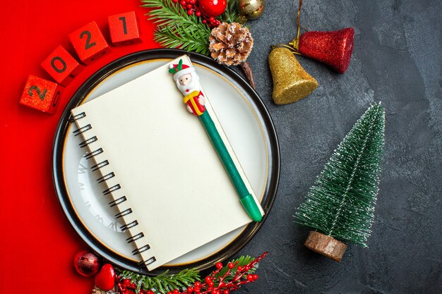 Visão aérea do fundo do ano novo com o caderno com a caneta no jantar acessórios de decoração ramos de abeto e números em um guardanapo vermelho ao lado da árvore de Natal em uma mesa preta