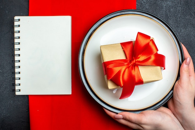 Visão aérea do plano de fundo da refeição de Natal nacional com a mão segurando pratos vazios com uma fita vermelha em forma de arco e um caderno em um guardanapo vermelho na mesa preta