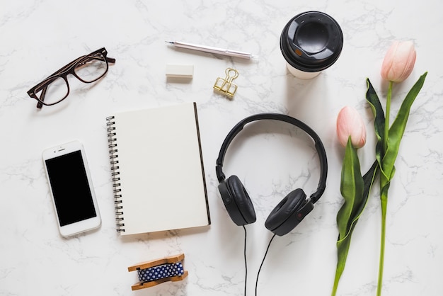 Visão aérea do telefone celular; Óculos; caderno; papelaria; fone de ouvido e tulipas cor de rosa no pano de fundo de mármore