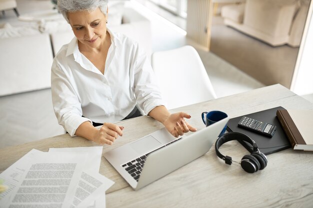 Visão de alto ângulo de uma elegante contadora madura confiante usando um laptop genérico para trabalho on-line, mantendo registros financeiros de grandes empresas, sentada à mesa com papéis