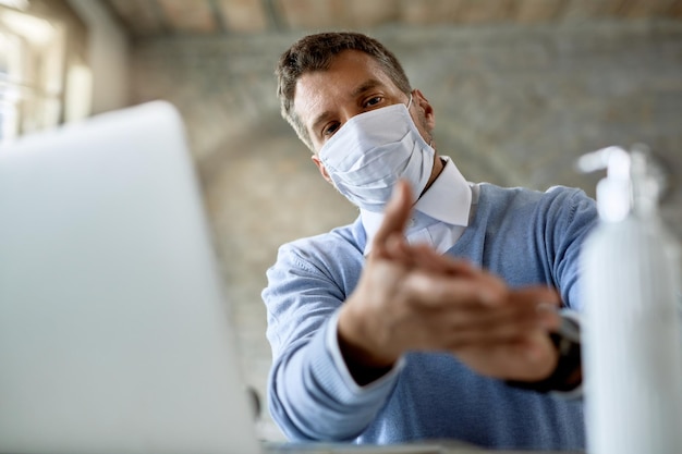Visão de ângulo baixo de um empresário com máscara facial protetora usando gel de mão antibacteriano enquanto trabalhava no escritório